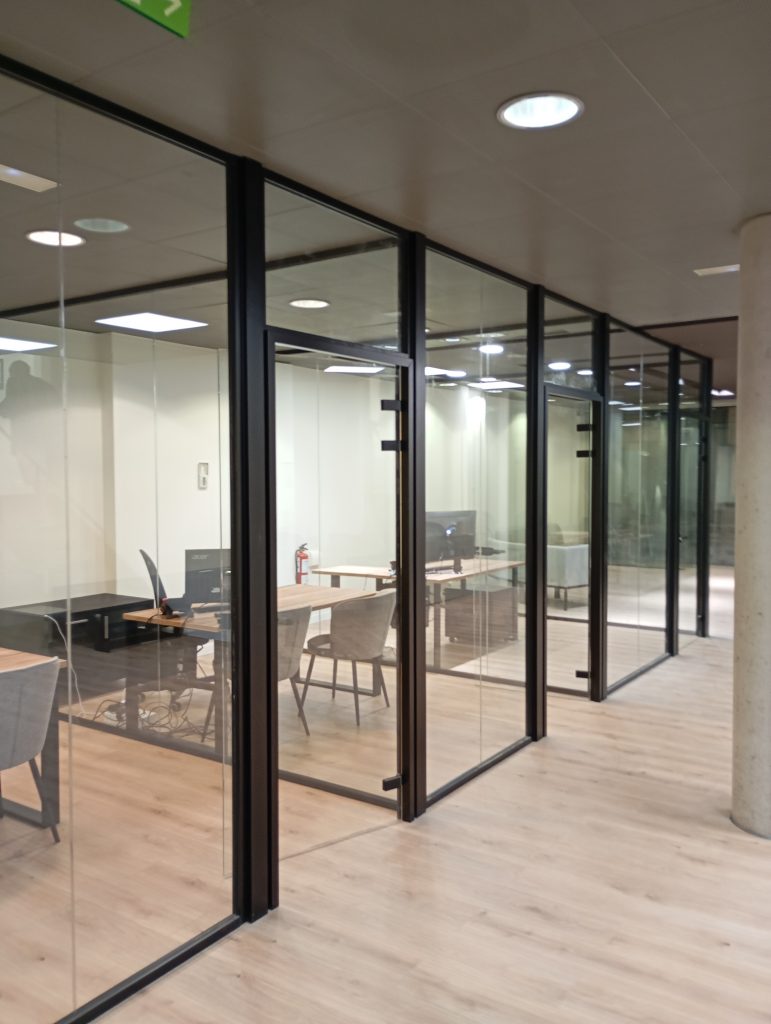 Mamparas de cristal con perfilería negra en oficina moderna, con despachos acristalados y suelo de madera clara.